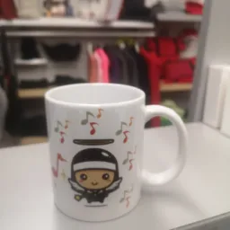 Biały kubek z nadrukiem postaci anioła-muzyka na tle rozmazanych ubrań w sklepie.