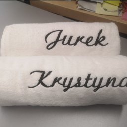 CustoMove - Dwa białe, puszyste ręczniki ułożone jeden na drugim, z wyhaftowanymi, czarnymi imionami: 'Jurek' na górnym i 'Krystyna' na dolnym. Ręczniki leżą na szarym blacie, w tle widoczny fragment biurka...