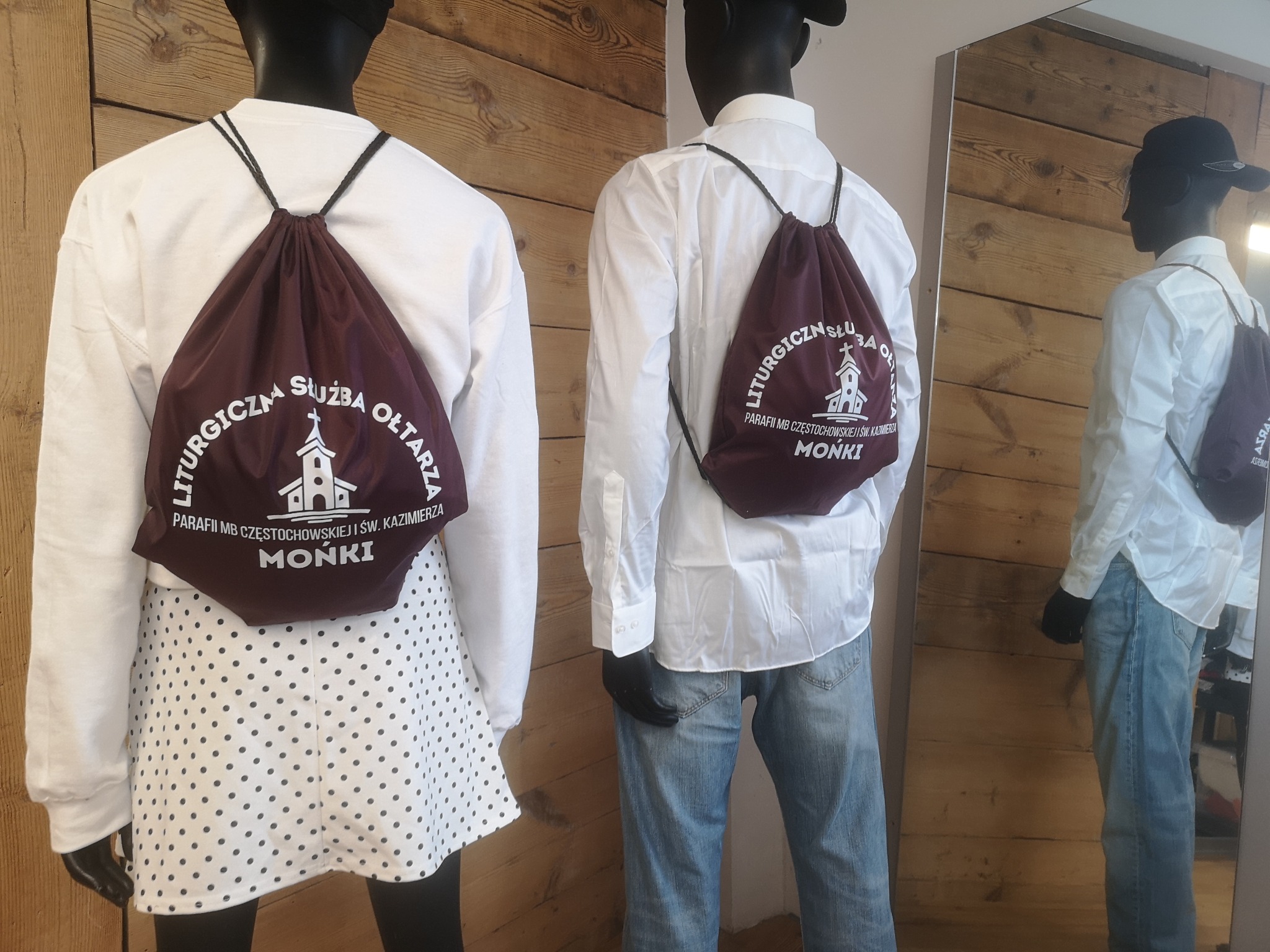 Dwa manekiny prezentują bordowe workoplecaki z białym nadrukiem 'Liturgiczna Służba Ołtarza Parafii MB Częstochowskiej i św. Kazimierza Mońki', jeden z manekinów ubrany w białą bluzę i spódnicę...