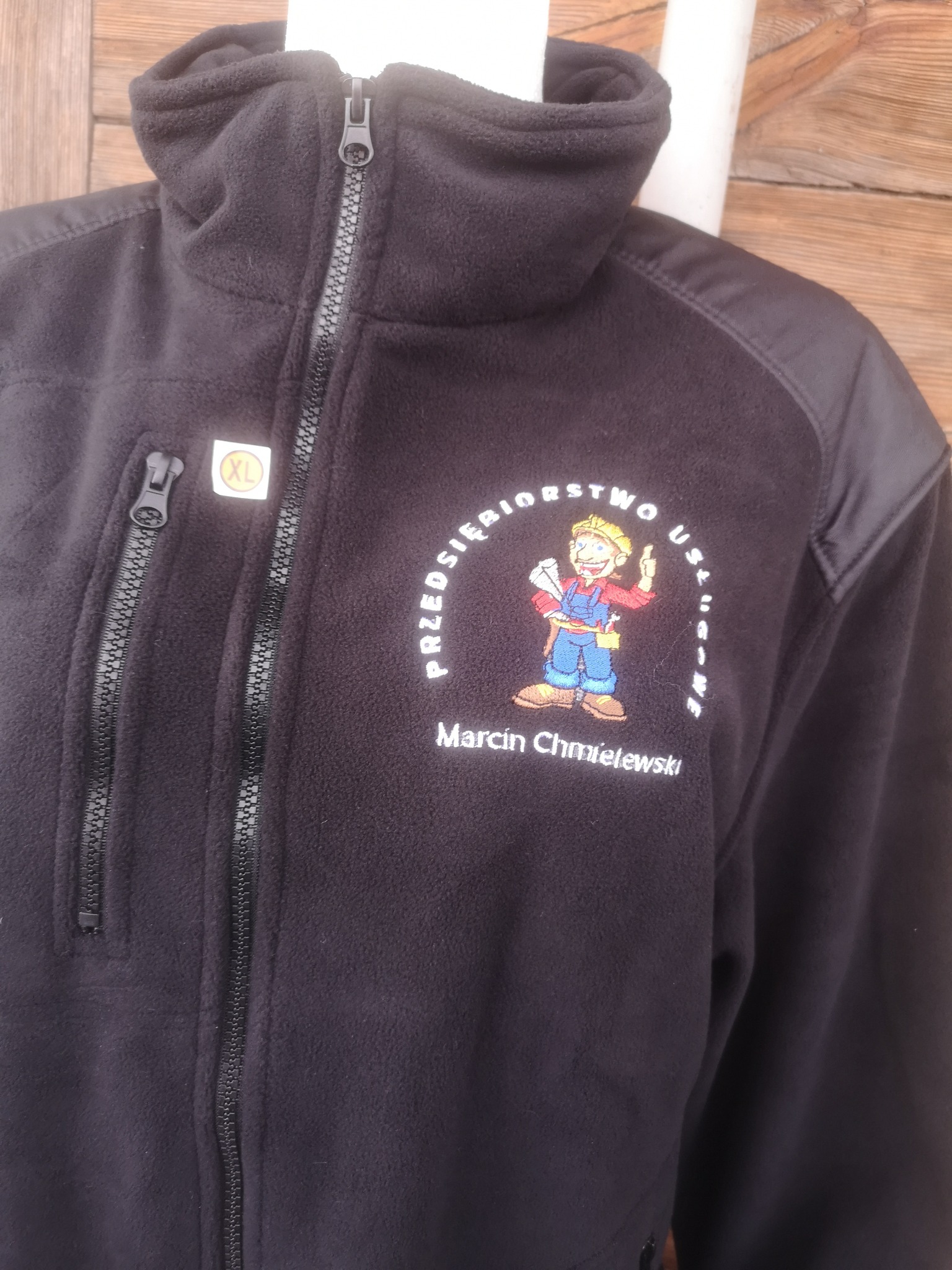 Czarna bluza polarowa z haftowanym logo firmy 'Przedsiębiorstwo Usługowe Marcin Chmielewski', przedstawiającym postać w kasku i kombinezonie. Bluza ma zamek błyskawiczny i kieszeń na zamek.