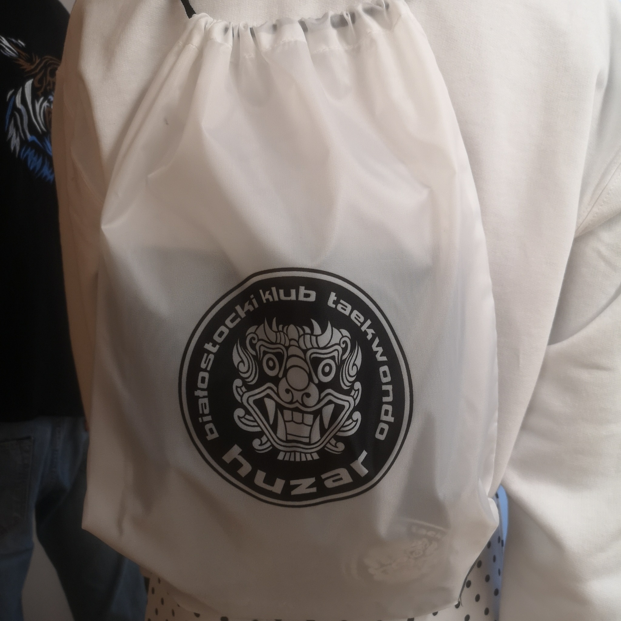 Biały worek sportowy z nadrukiem logo Białostockiego Klubu Taekwondo Huzar noszony na plecach osoby w białej bluzie.