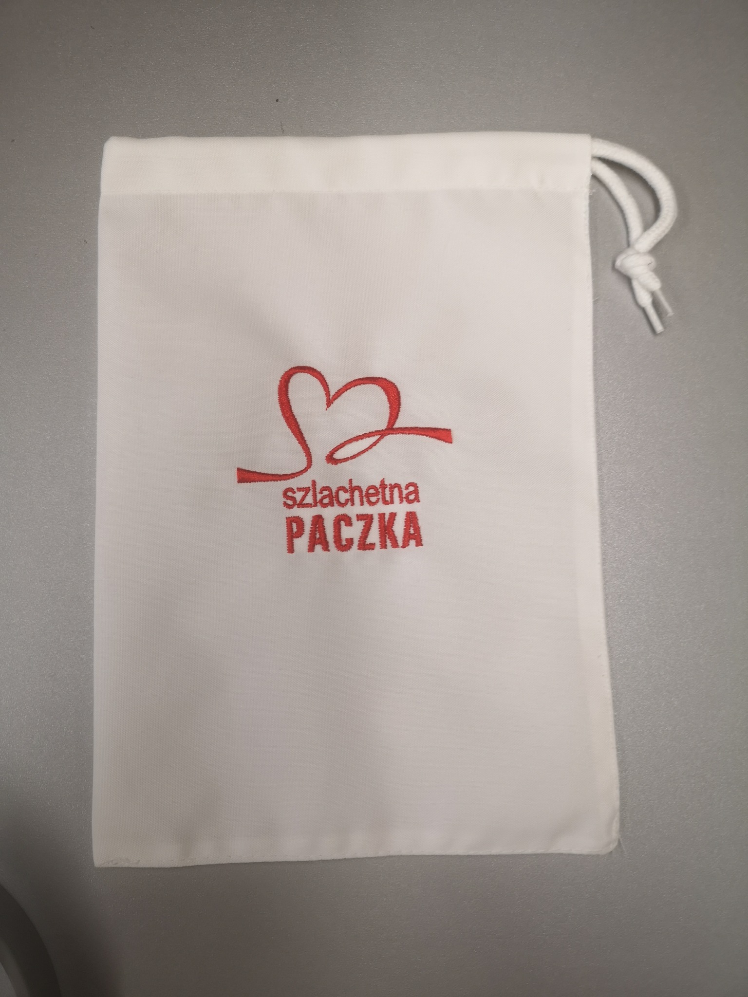 Biały woreczek ze sznurkiem, z czerwonym haftowanym logo 'Szlachetna Paczka', leżący na szarym tle.