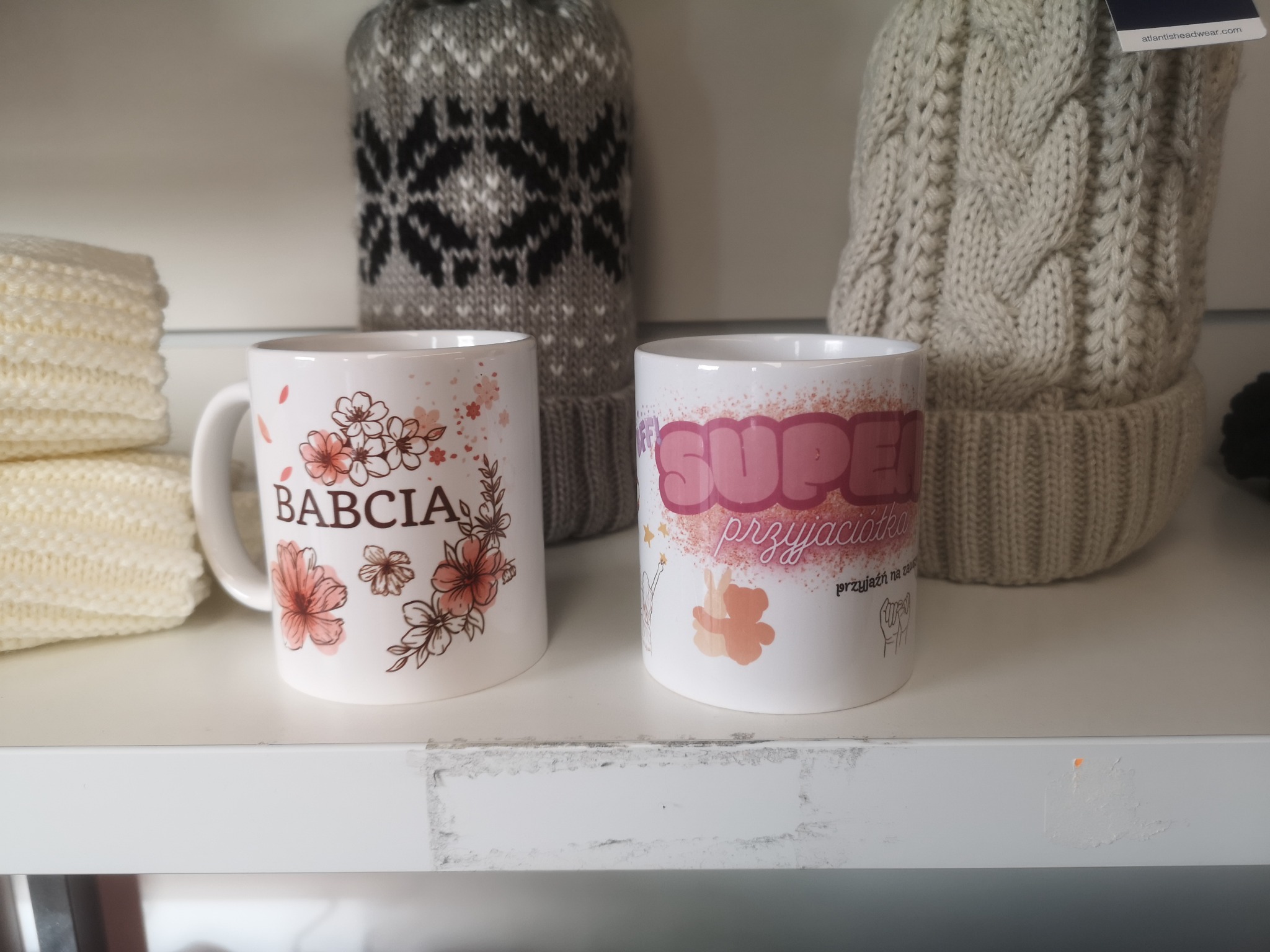 Dwa białe kubki z różnymi nadrukami (kwiaty i napis 'Babcia' oraz napis 'Super Przyjaciółka') stoją na białej półce, w tle widać dwie wełniane czapki.