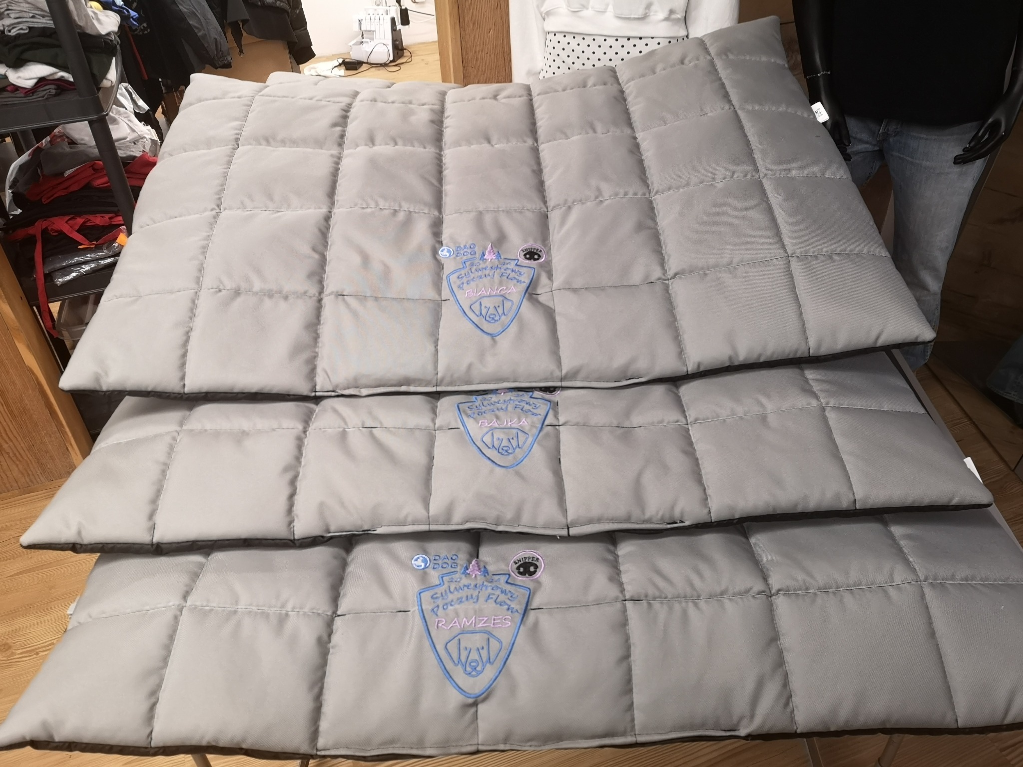 Stos trzech szarych, pikowanych mat z wyhaftowanym logo firmy dla psów, każda z innym imieniem: Bajka, Ramzes, Bianca.