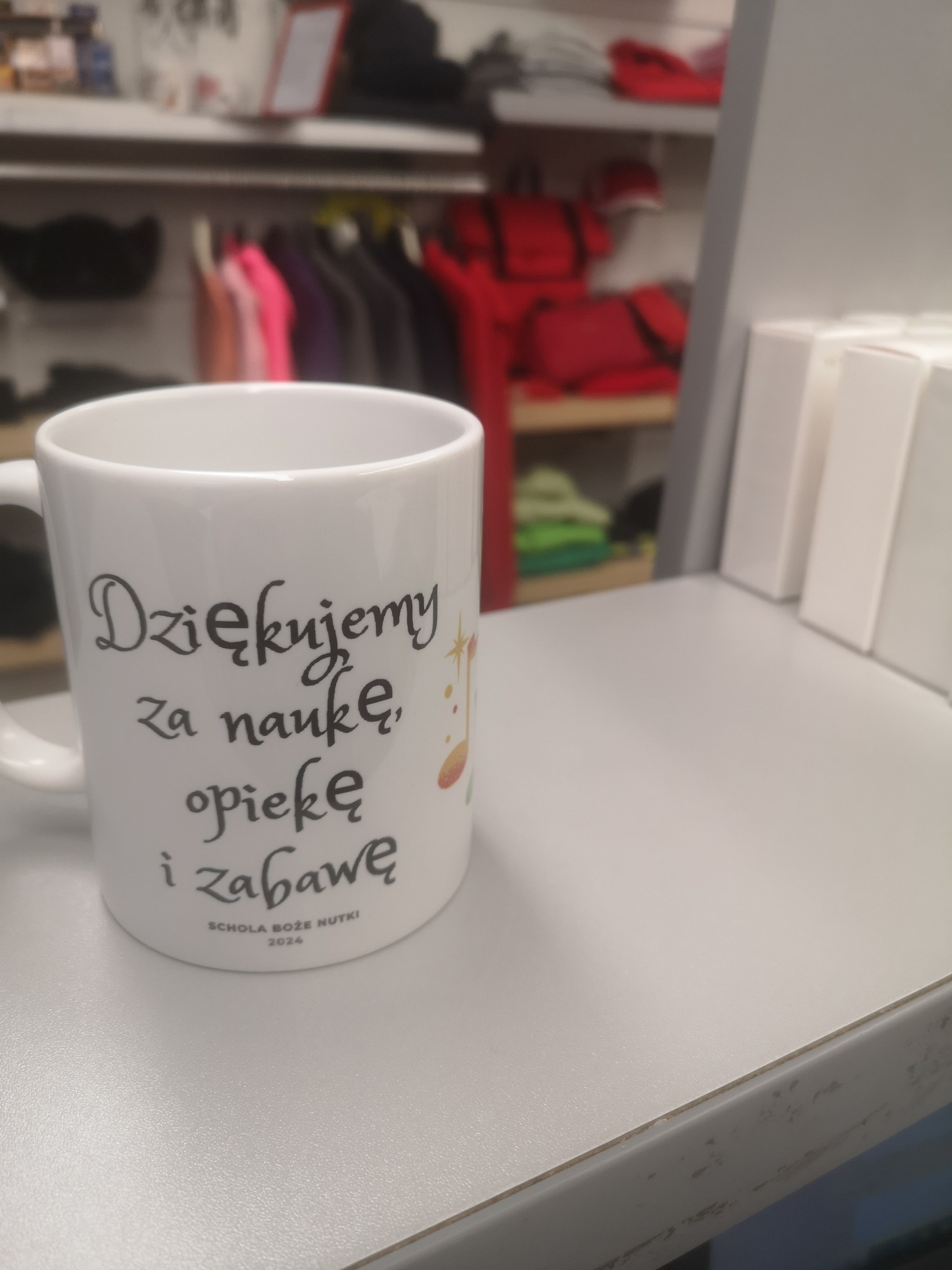 Biały kubek z czarnym napisem 'Dziękujemy za naukę, opiekę i zabawę' oraz nazwą szkoły 'SCHOLA BOŻE NUTKI 2024', stojący na jasnym blacie na tle wieszaków z odzieżą.
