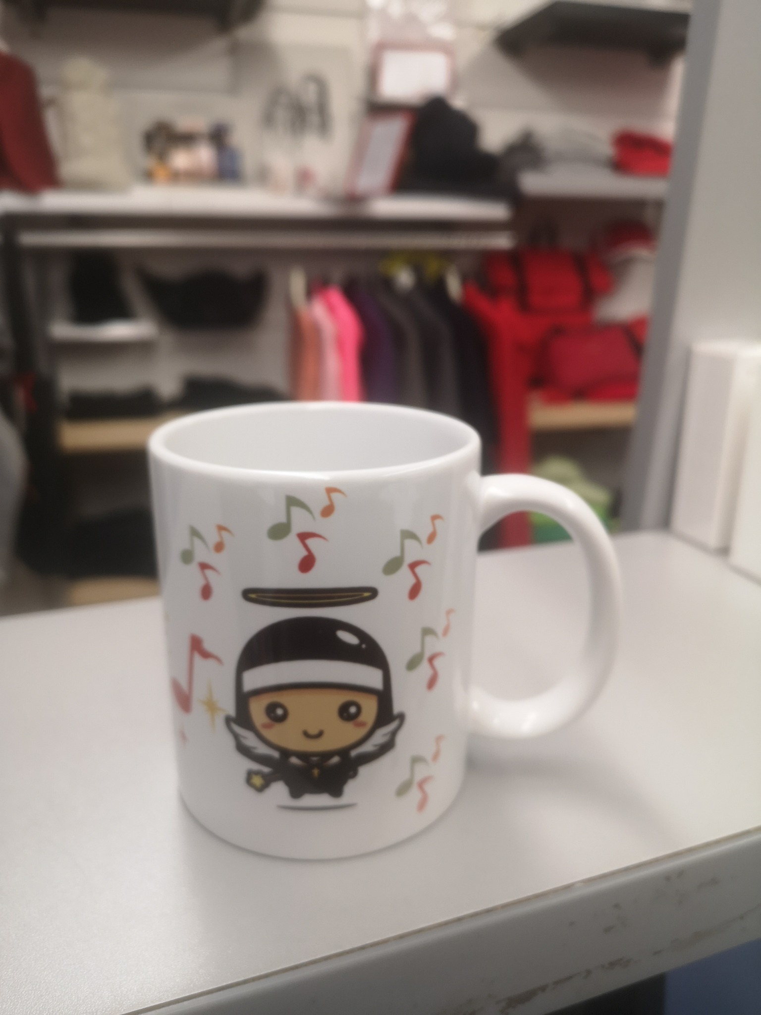 Biały kubek z nadrukiem postaci anioła-muzyka na tle rozmazanych ubrań w sklepie.