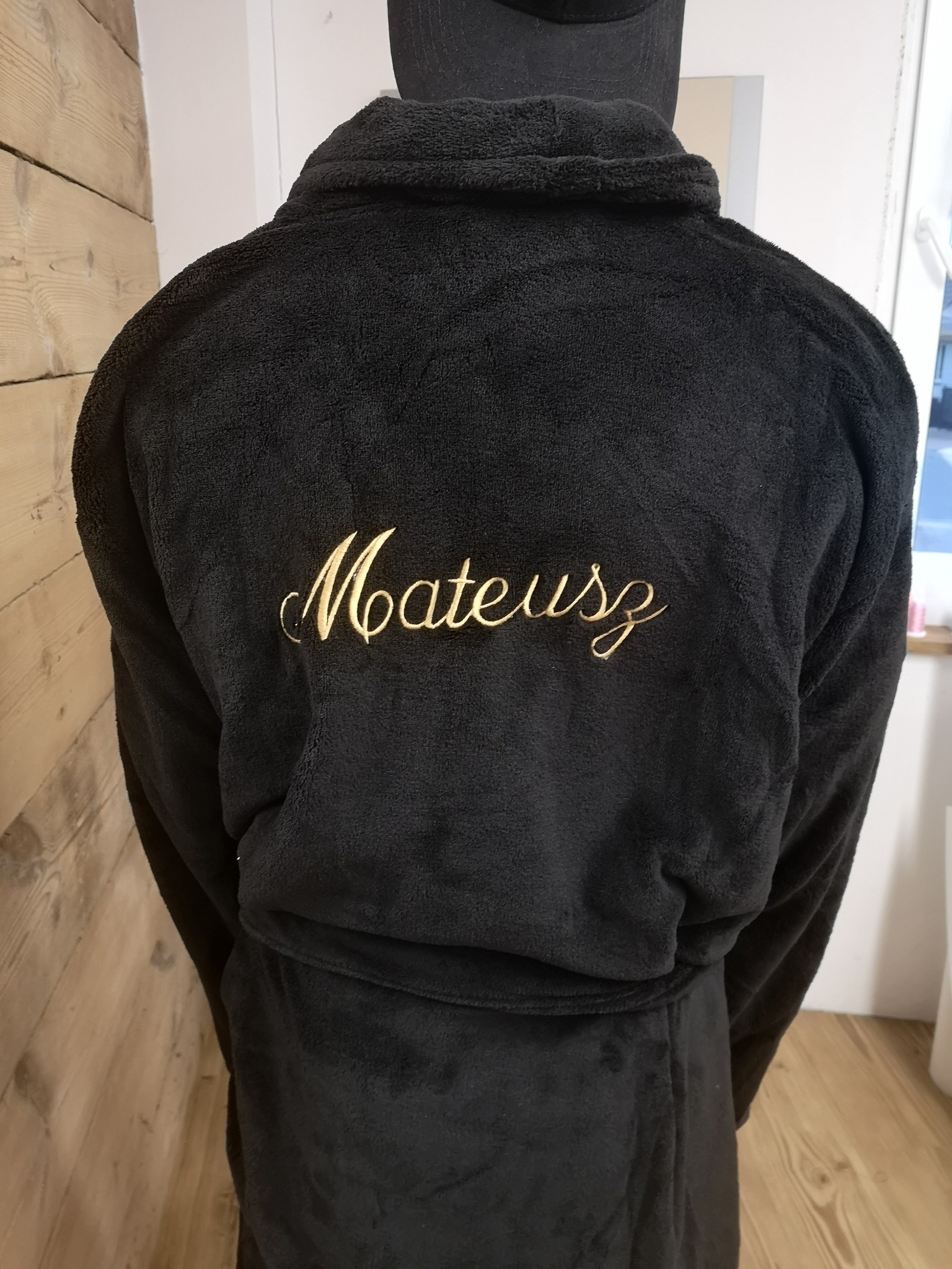 Czarny, pluszowy szlafrok z wyhaftowanym złotą nicią imieniem 'Mateusz' na plecach, widok z tyłu na osobę w szlafroku.