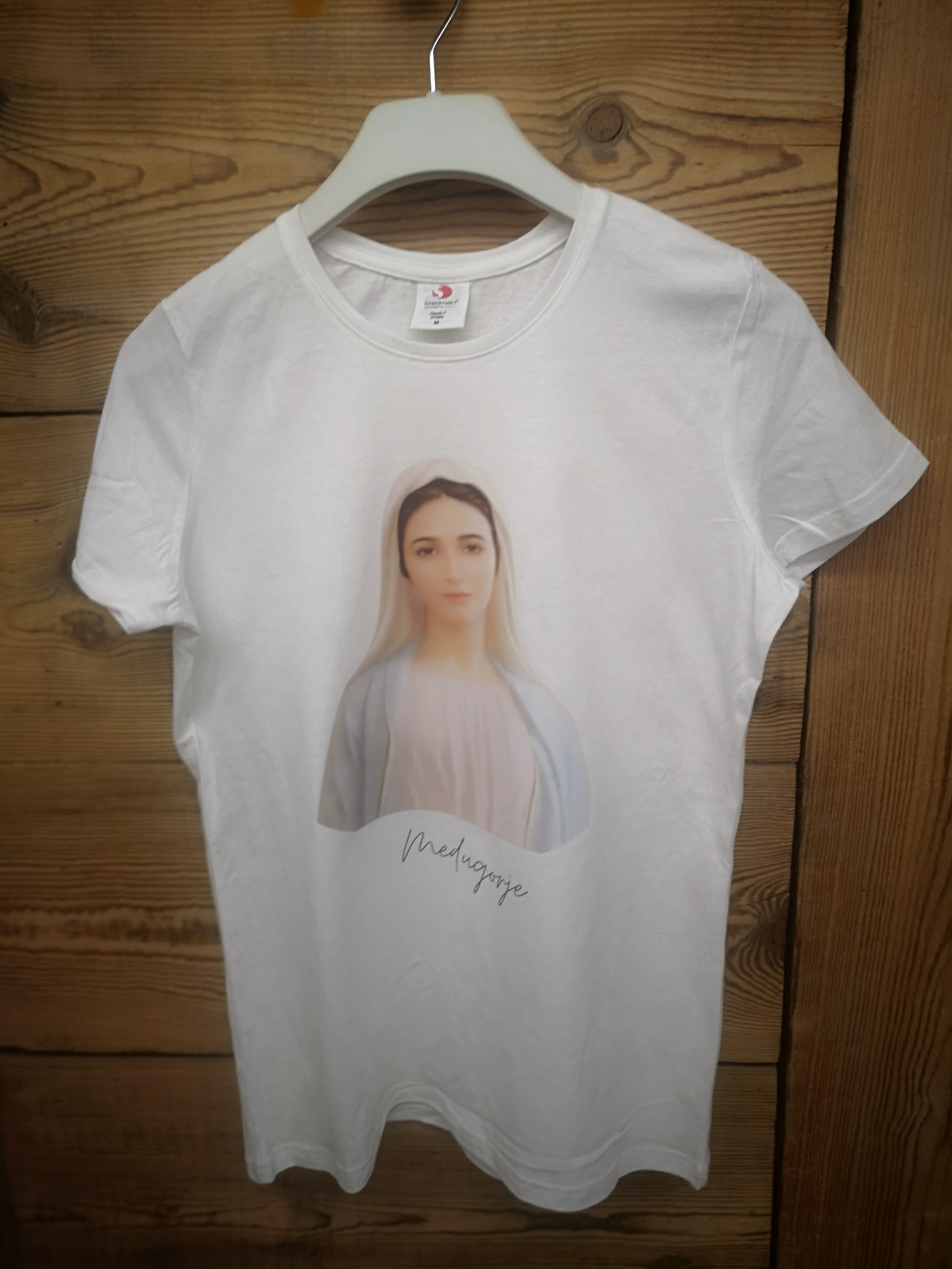 Biała koszulka z nadrukiem wizerunku Matki Bożej z Medjugorie, wisząca na wieszaku na tle drewnianej ściany.