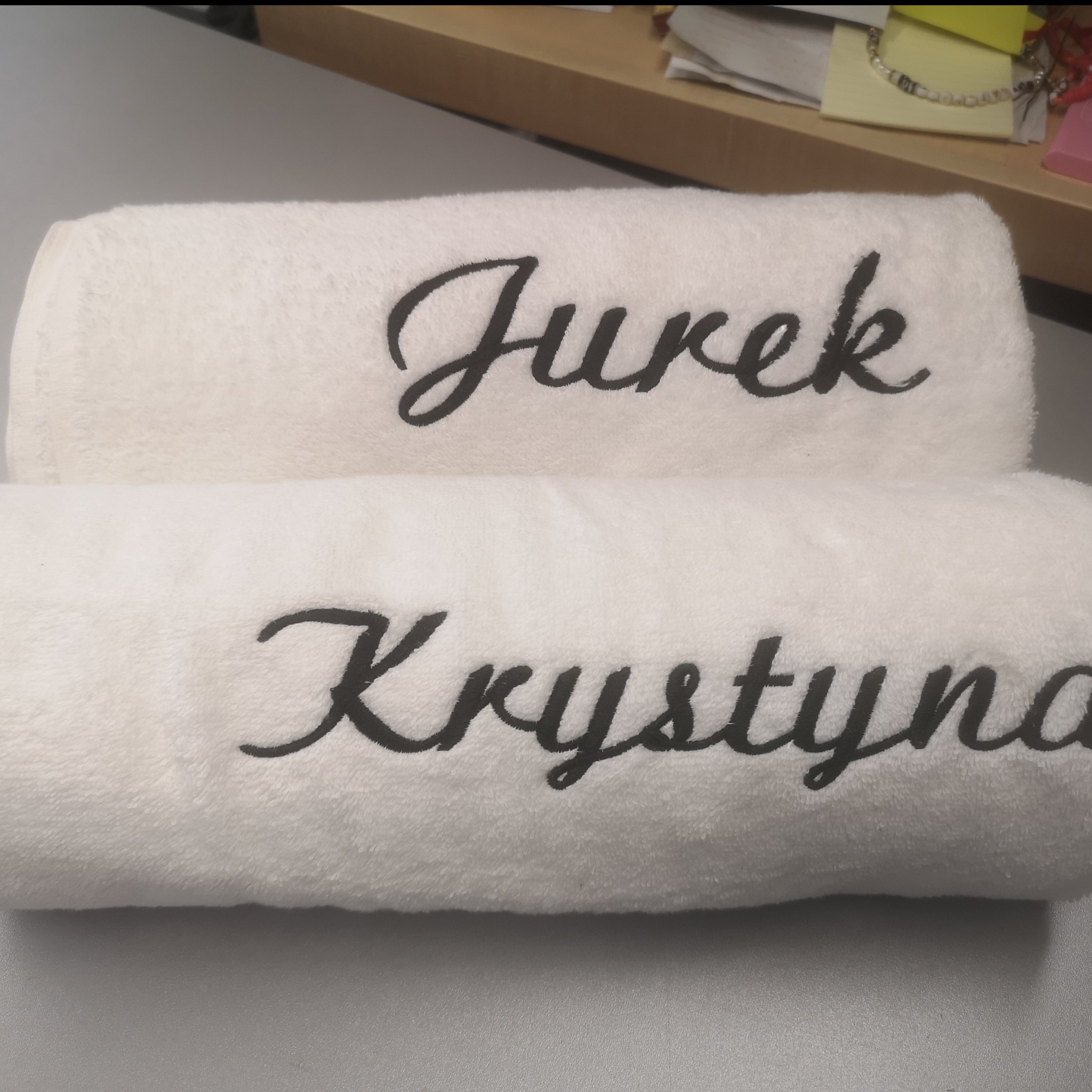 Dwa białe, puszyste ręczniki ułożone jeden na drugim, z wyhaftowanymi, czarnymi imionami: 'Jurek' na górnym i 'Krystyna' na dolnym. Ręczniki leżą na szarym blacie, w tle widoczny fragment biurka...