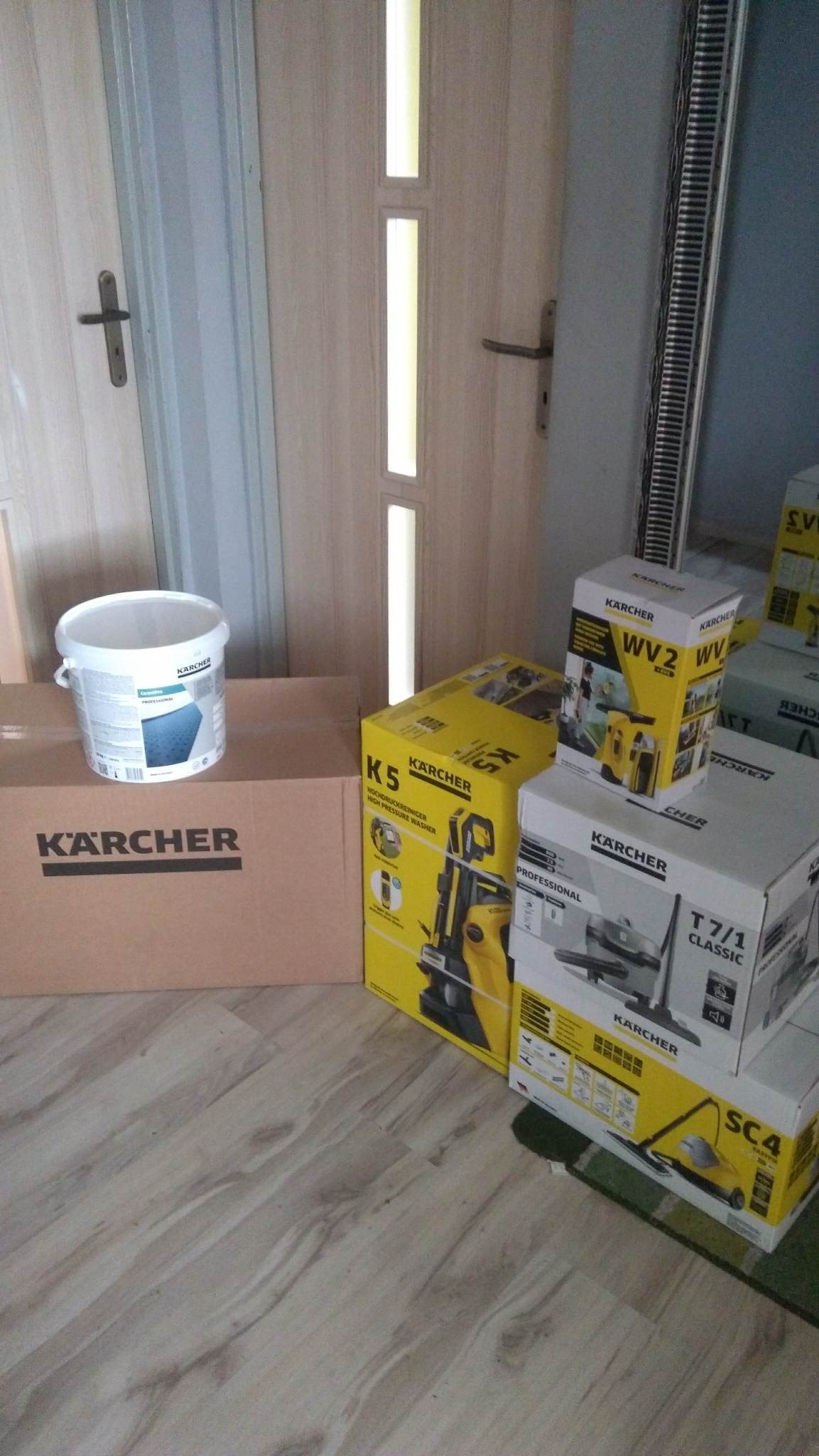 Pudełka i pojemnik z produktami marki Kärcher: myjka ciśnieniowa K5, odkurzacz do okien WV2, odkurzacz T 7/1 Classic, parownica SC4 oraz środek czyszczący w białym wiaderku, ustawione na podłodze...