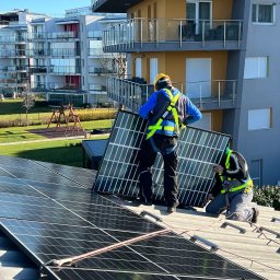 MS SOLAR ENERGY Sp. z o.o. - Dwóch instalatorów w uprzężach zabezpieczających montuje panel słoneczny na dachu budynku mieszkalnego; w tle widoczne kolejne budynki i plac zabaw.