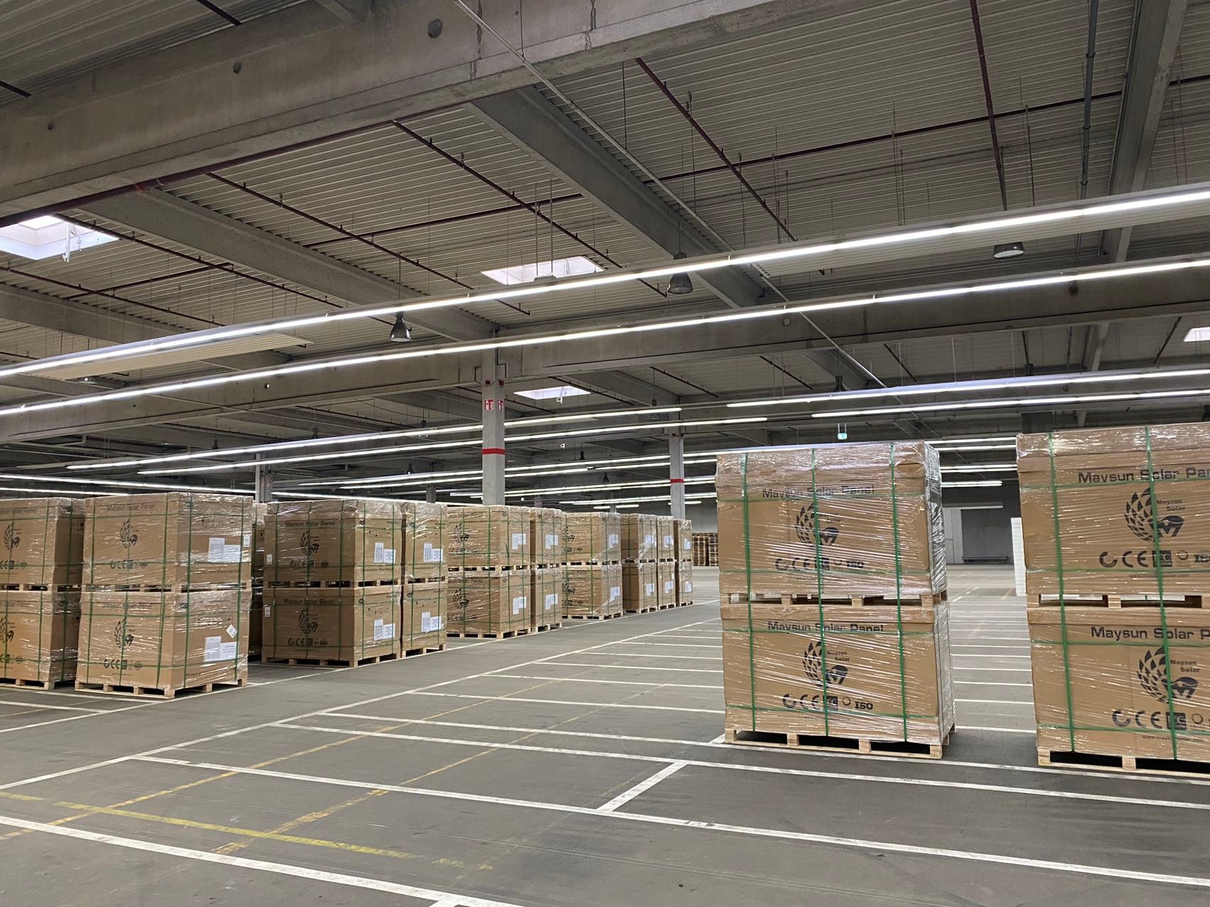 Magazyn w Hilden, Niemcy, wypełniony paletami z panelami słonecznymi Maysun Solar, zabezpieczonymi folią i zielonymi taśmami, widoczne oświetlenie LED pod sufitem