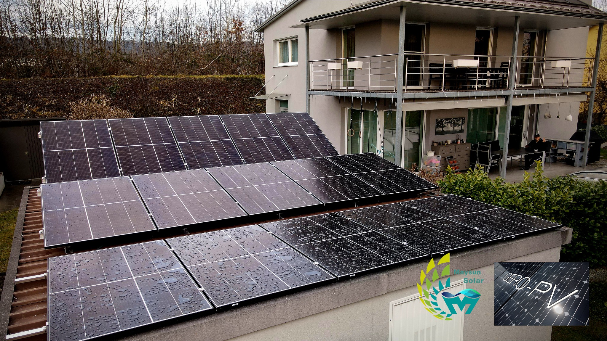 Panele słoneczne Maysun Solar 410W zamontowane na dachu dwukondygnacyjnego domu, widoczne krople wody na powierzchni, w tle drzewa bez liści i osoba siedząca na tarasie. W prawym dolnym rogu logo...