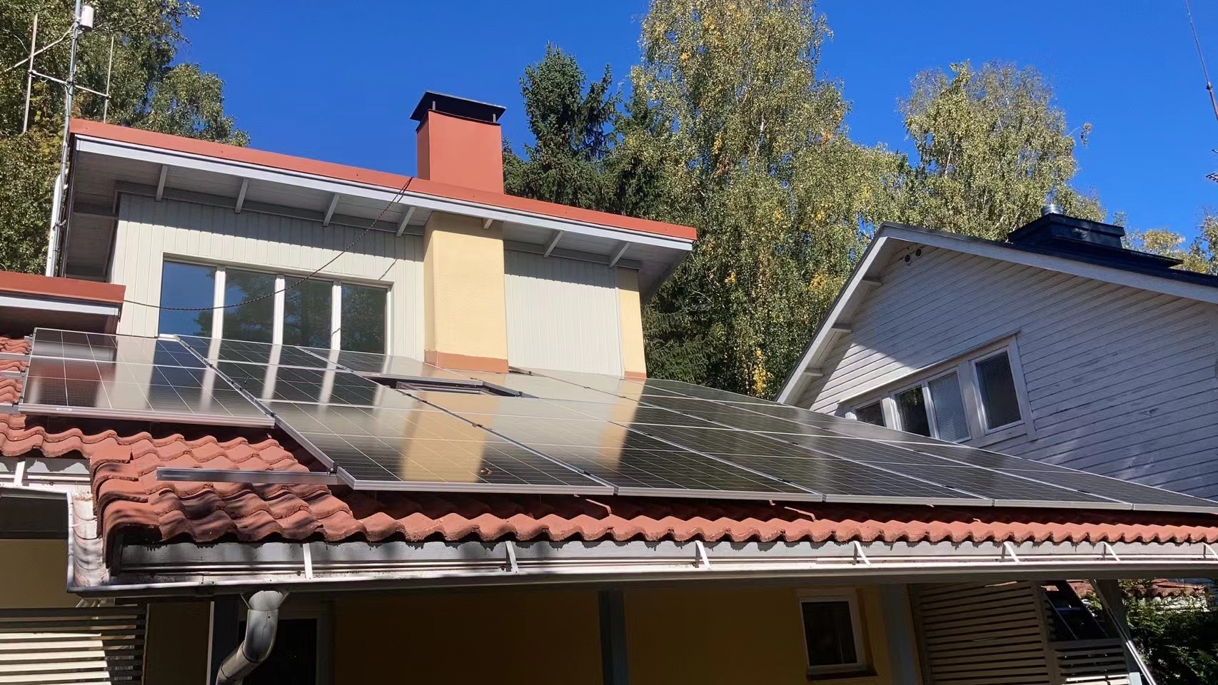 Panele fotowoltaiczne zamontowane na dachu domu jednorodzinnego z czerwoną dachówką, widoczne okna i komin, błękitne niebo w tle.