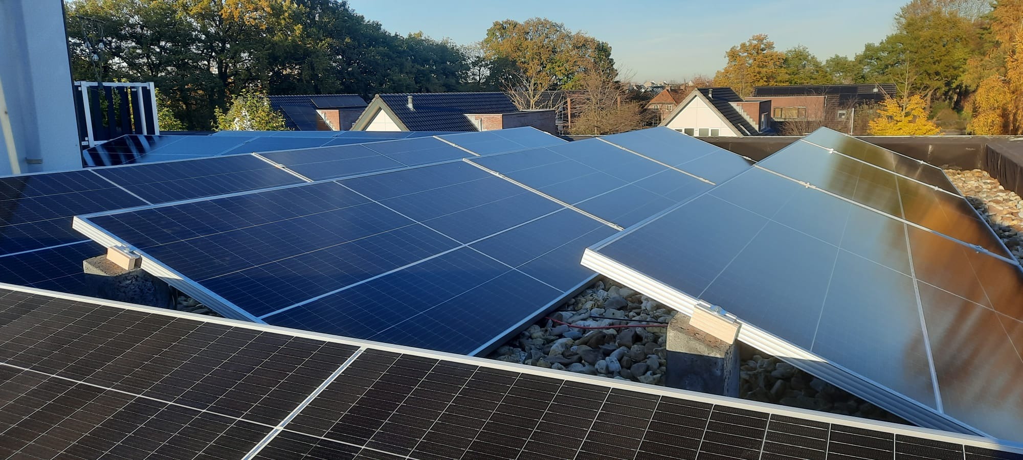 Instalacja paneli słonecznych na płaskim dachu, widoczne panele Maysun Solar 510W, podtrzymywane na betonowych blokach i otoczone drobnymi kamieniami, w tle zabudowa jednorodzinna i drzewa.