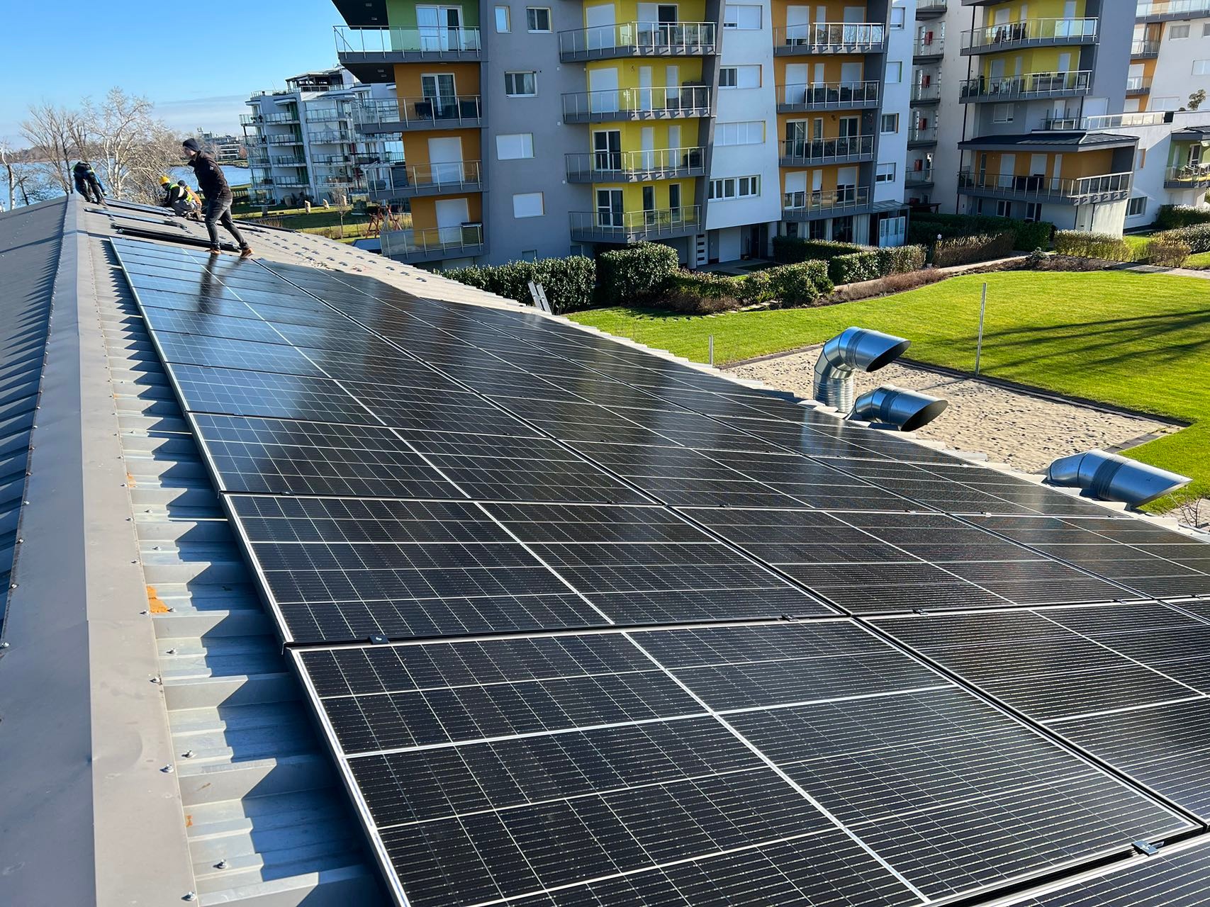 Instalacja paneli słonecznych na dachu budynku mieszkalnego w słoneczny dzień. Widoczni pracownicy w trakcie montażu, panele Maysun Solar oraz elementy wentylacji dachowej. W tle budynek mieszkalny...
