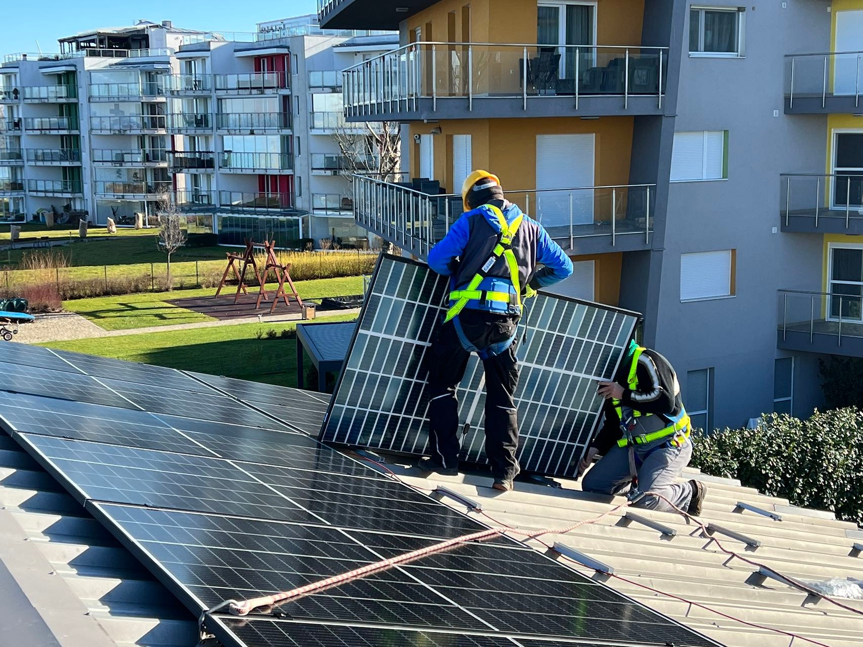 Dwóch instalatorów w uprzężach zabezpieczających montuje panel słoneczny na dachu budynku mieszkalnego; w tle widoczne kolejne budynki i plac zabaw.