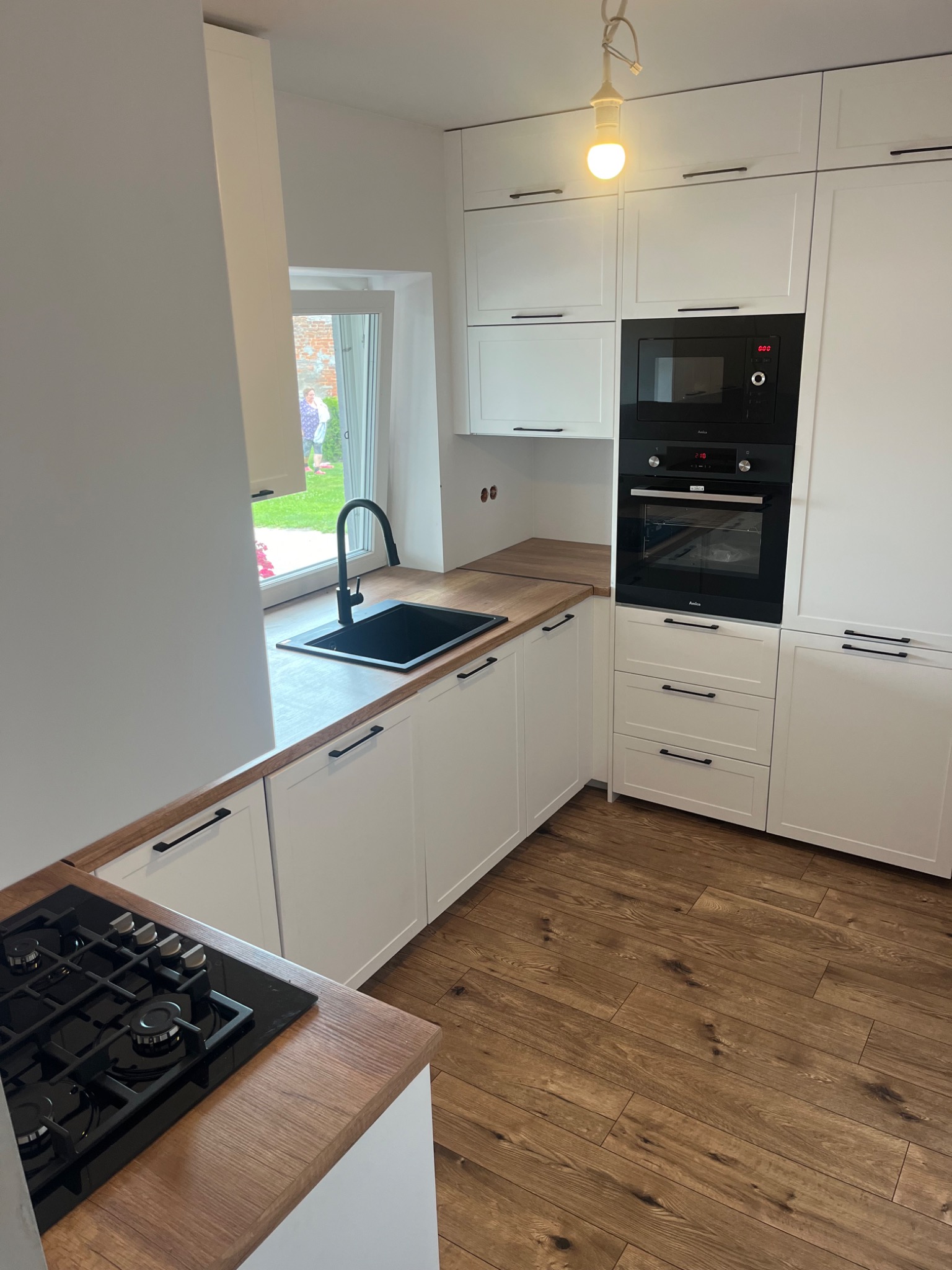 Nowoczesna kuchnia z białymi szafkami, drewnianym blatem i czarnymi akcentami, w tym zlewozmywakiem, baterią i płytą grzewczą, z widokiem na ogród przez okno.