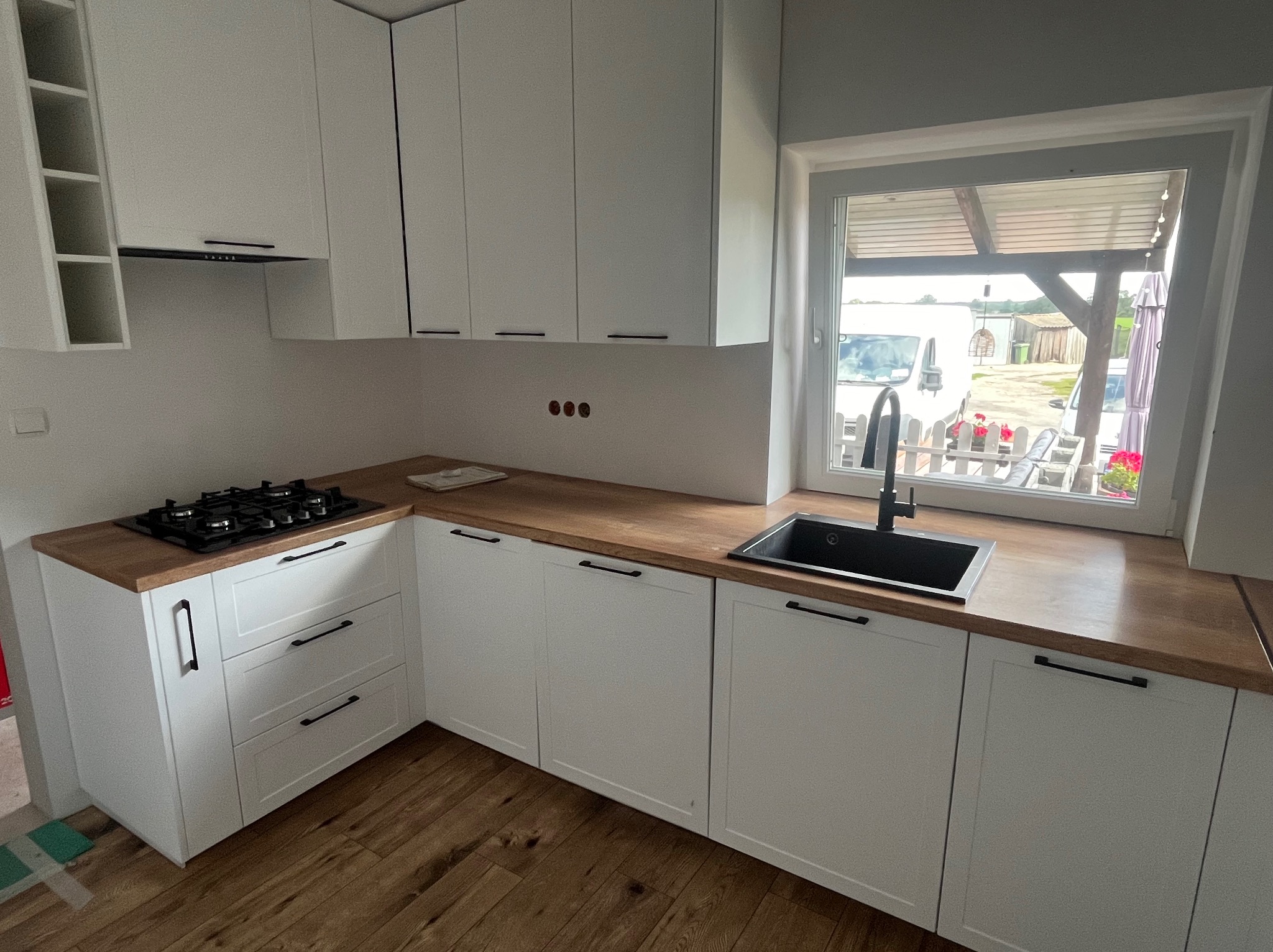 Nowoczesna, jasna kuchnia z białymi szafkami i drewnianym blatem, czarną armaturą i płytą gazową. Widok na zewnątrz przez okno.