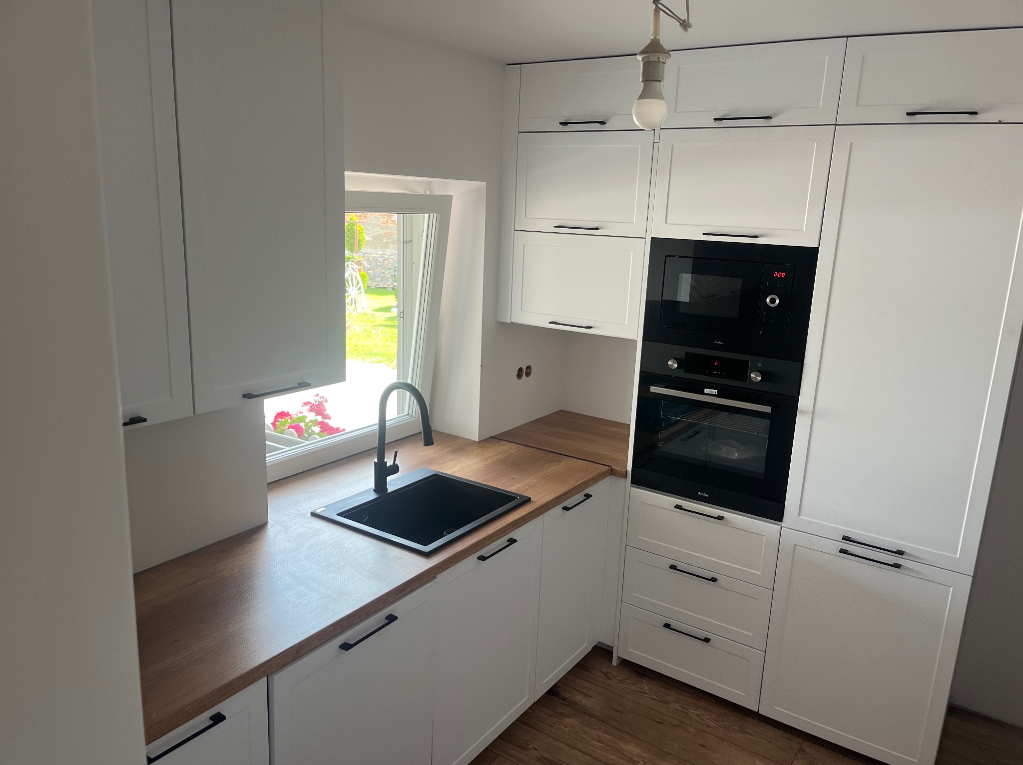 Nowoczesna, biała kuchnia z drewnianym blatem, czarnym zlewem i armaturą. W tle okno z widokiem na ogród.