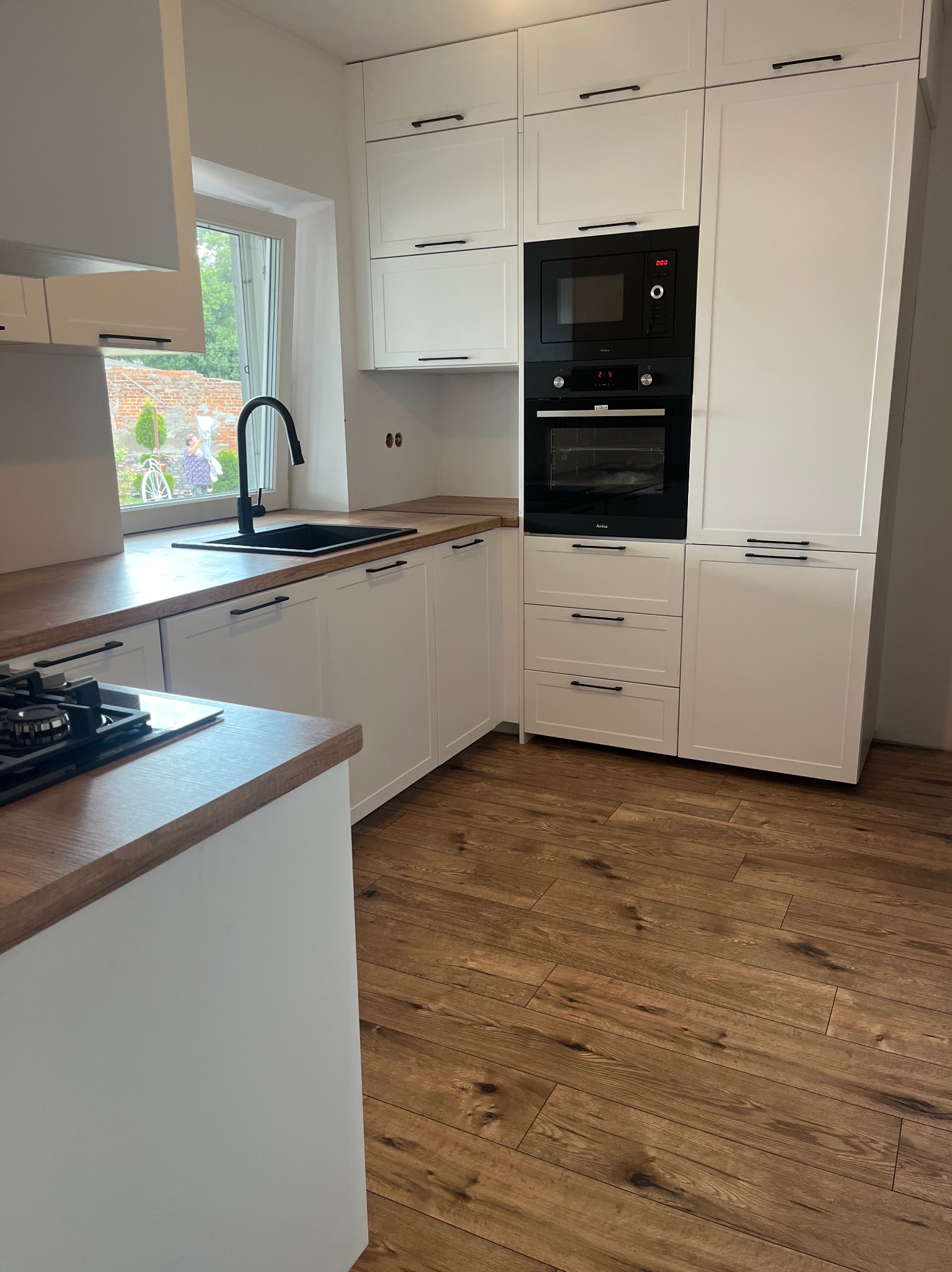 Nowoczesna, biała kuchnia z drewnianym blatem w Starym Mieście. Widoczne czarne, minimalistyczne uchwyty, piekarnik i mikrofalówka w zabudowie oraz czarny zlew z baterią. Za oknem widoczny ogród...