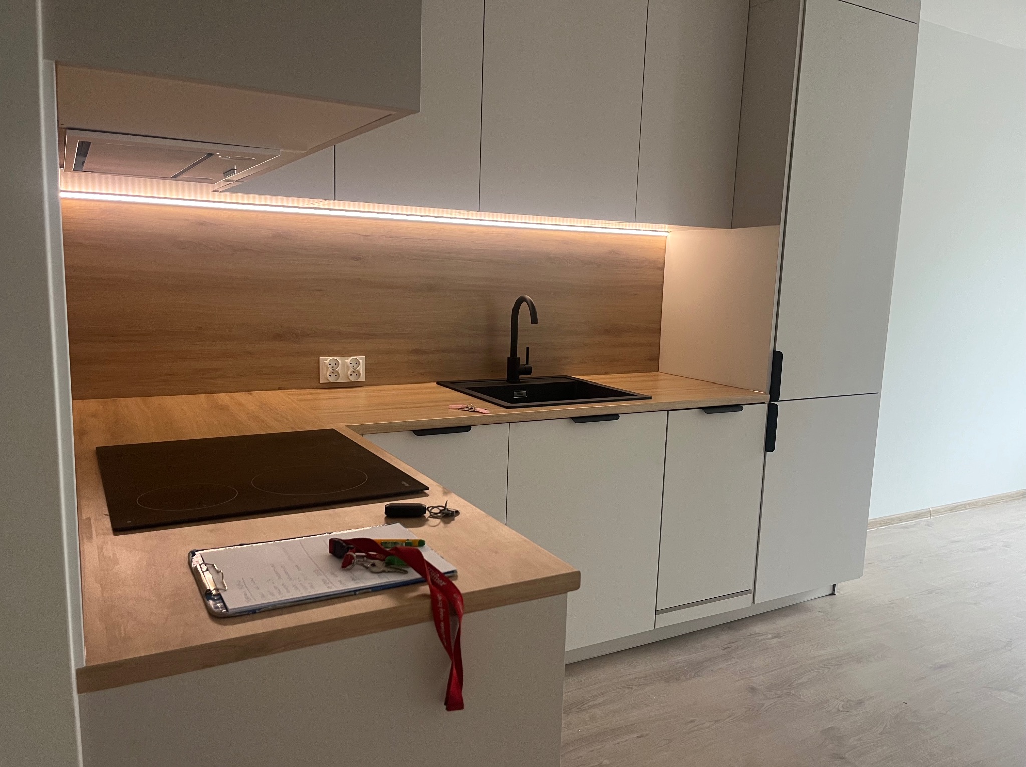 Nowoczesna kuchnia z białymi szafkami, drewnianym blatem i czarną armaturą. Na blacie leży notatnik z długopisem i kluczami.