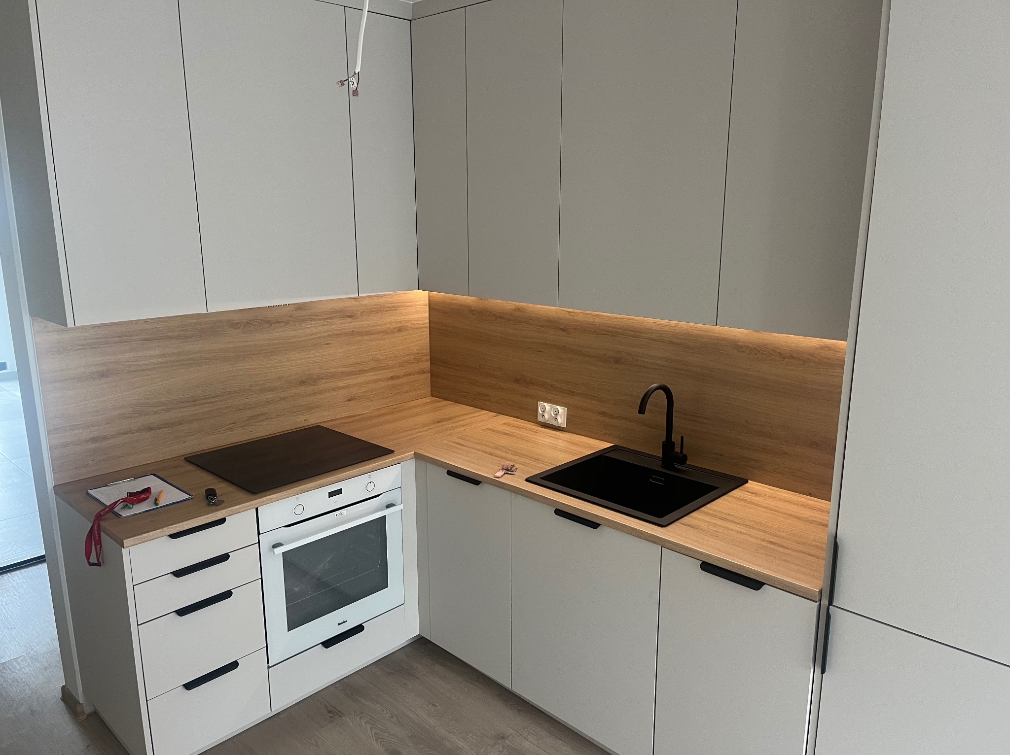 Nowoczesna kuchnia z szarymi szafkami, drewnianym blatem i czarnym zlewem, minimalistyczny design, oświetlenie LED pod szafkami, widoczne gniazdka elektryczne na ścianie.