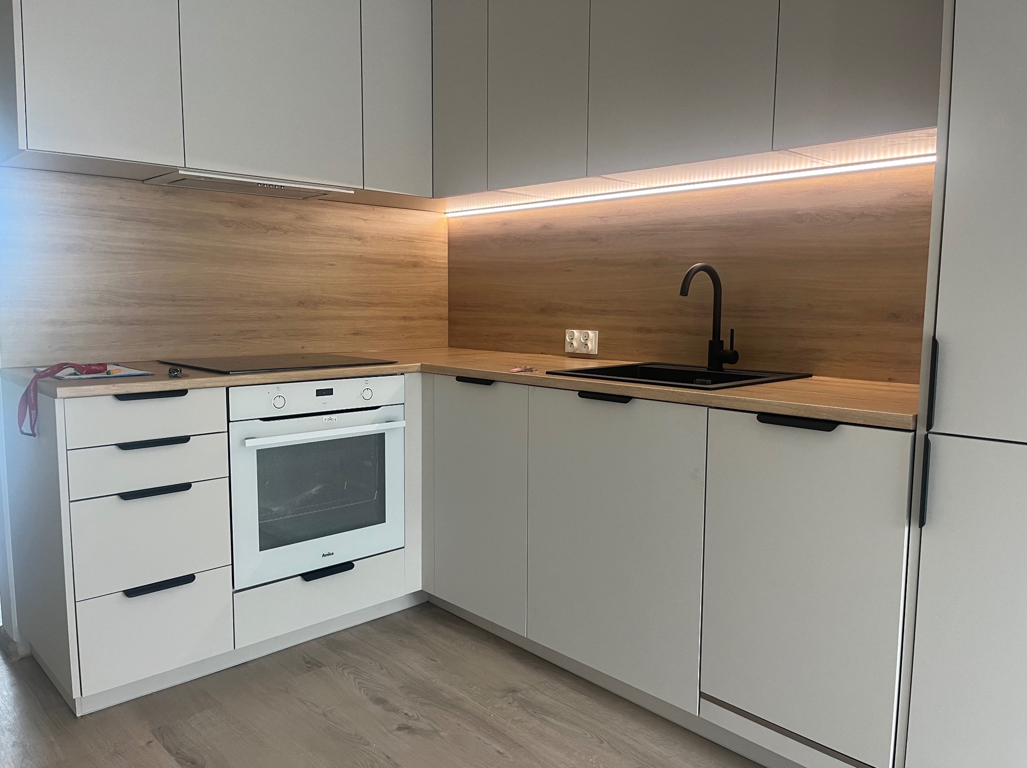 Nowoczesna kuchnia w odcieniach szarości i drewna z czarną armaturą, podświetleniem LED podszafkowym i sprzętem AGD marki Amica.