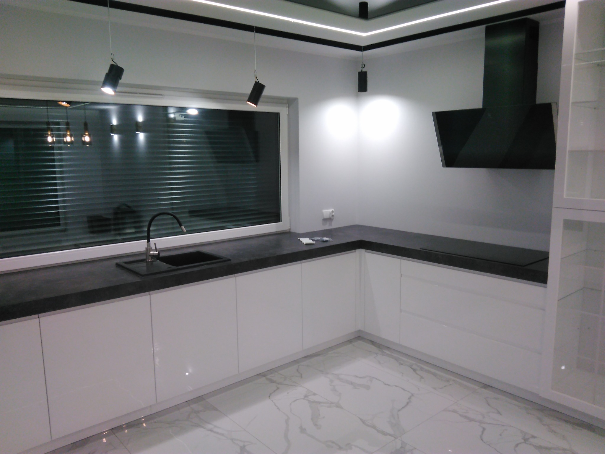 Nowoczesna kuchnia w bieli z czarnym blatem i okapem, oświetlenie punktowe, marmurowe płytki podłogowe.