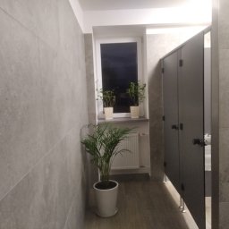 PETROBUD Petro Rakochyi - Wnętrze publicznej toalety z szarymi płytkami na ścianach, kabinami z ciemnego drewna i roślinami doniczkowymi przy oknie.