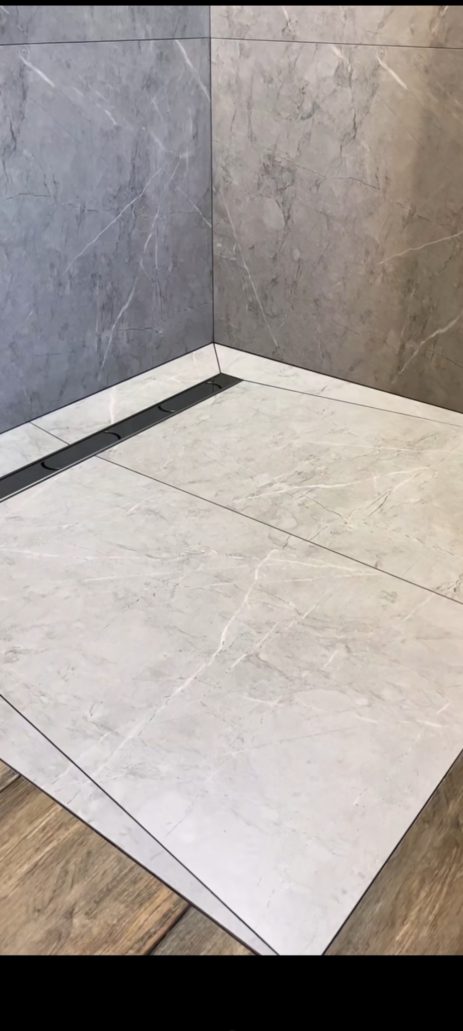 Wykończona kabina prysznicowa z szarymi płytkami imitującymi marmur na ścianach i podłodze, z czarnym odpływem liniowym i fragmentem drewnopodobnych płytek podłogowych na dole kadru.