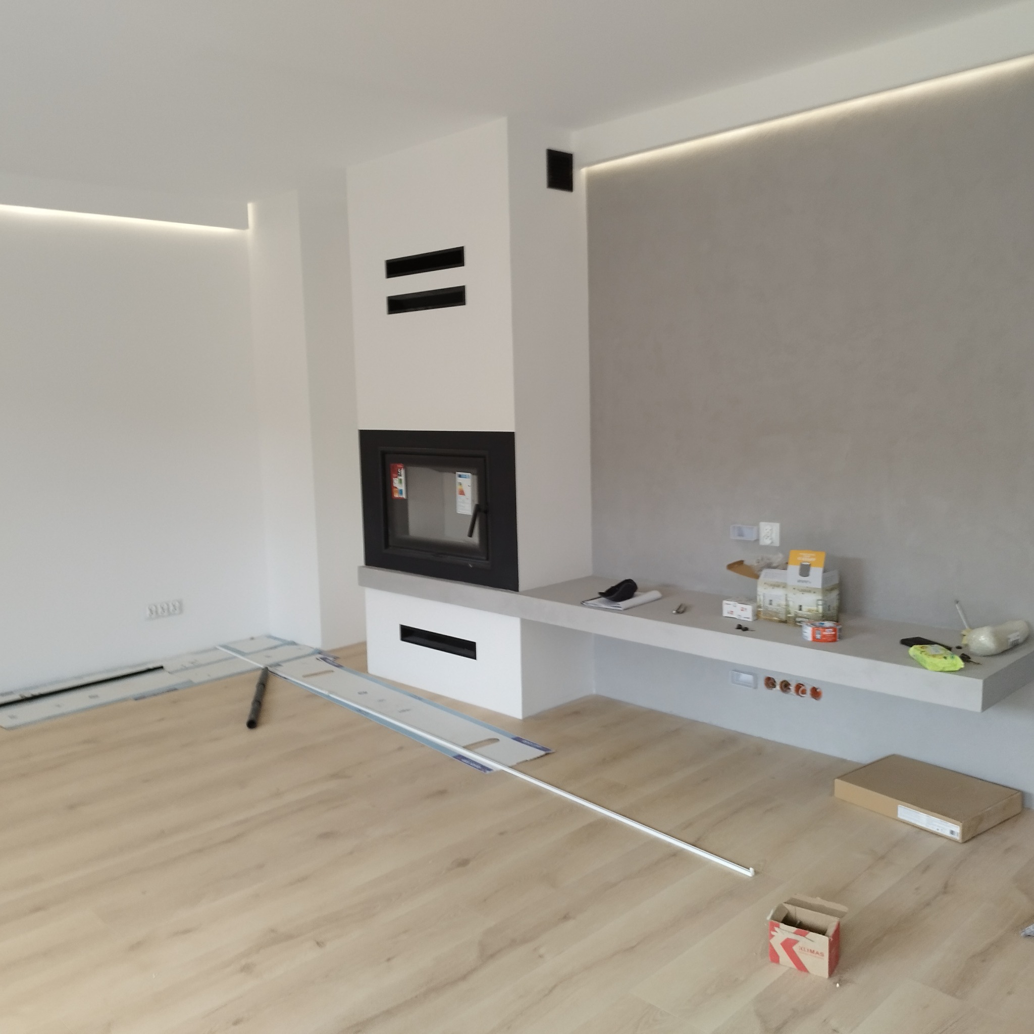 Salon w trakcie wykańczania: jasne panele podłogowe, białe ściany z ukrytym oświetleniem LED, szara ściana dekoracyjna, nowoczesny kominek z czarną obudową, narzędzia i materiały budowlane...