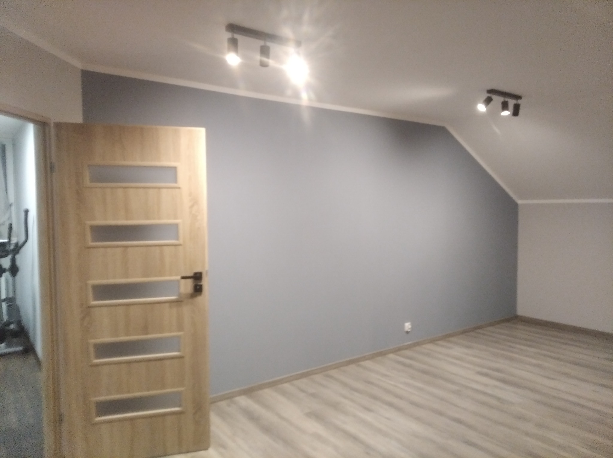 Jasne panele podłogowe w pokoju na poddaszu z szarą ścianą i drewnianymi drzwiami z przeszkleniami, oświetlenie punktowe sufitowe.