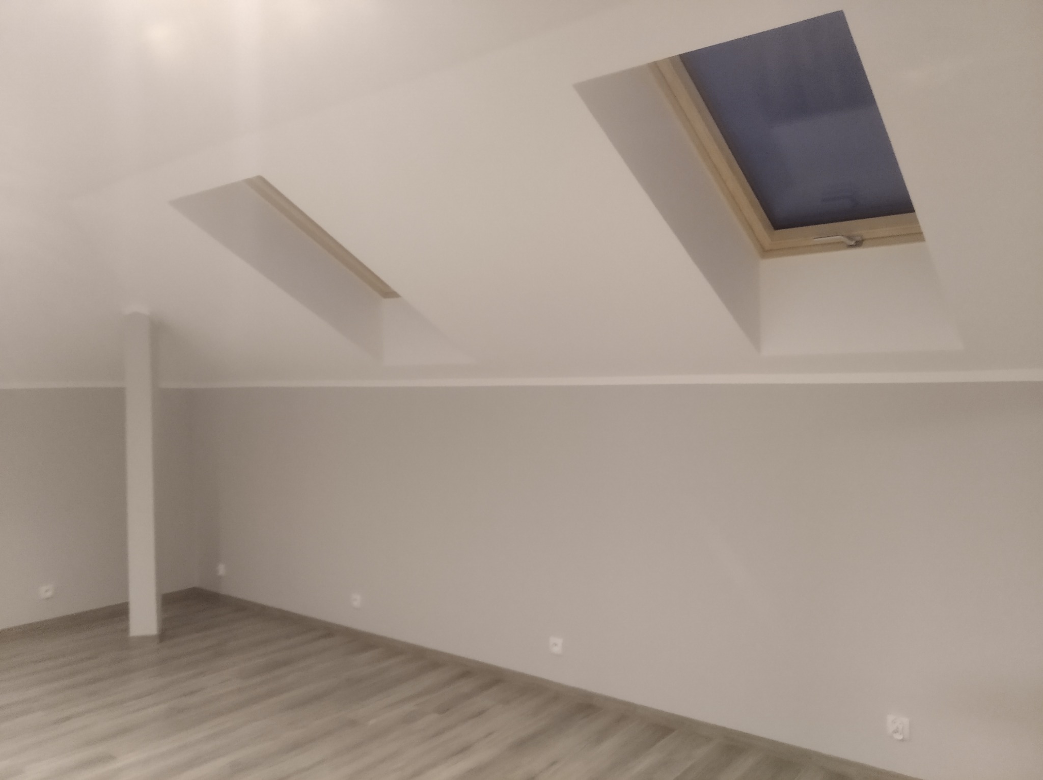 Poddasze po remoncie: gładkie, szare ściany, jasna podłoga z paneli, dwa okna dachowe z drewnianymi ramami, widoczny fragment słupa konstrukcyjnego.