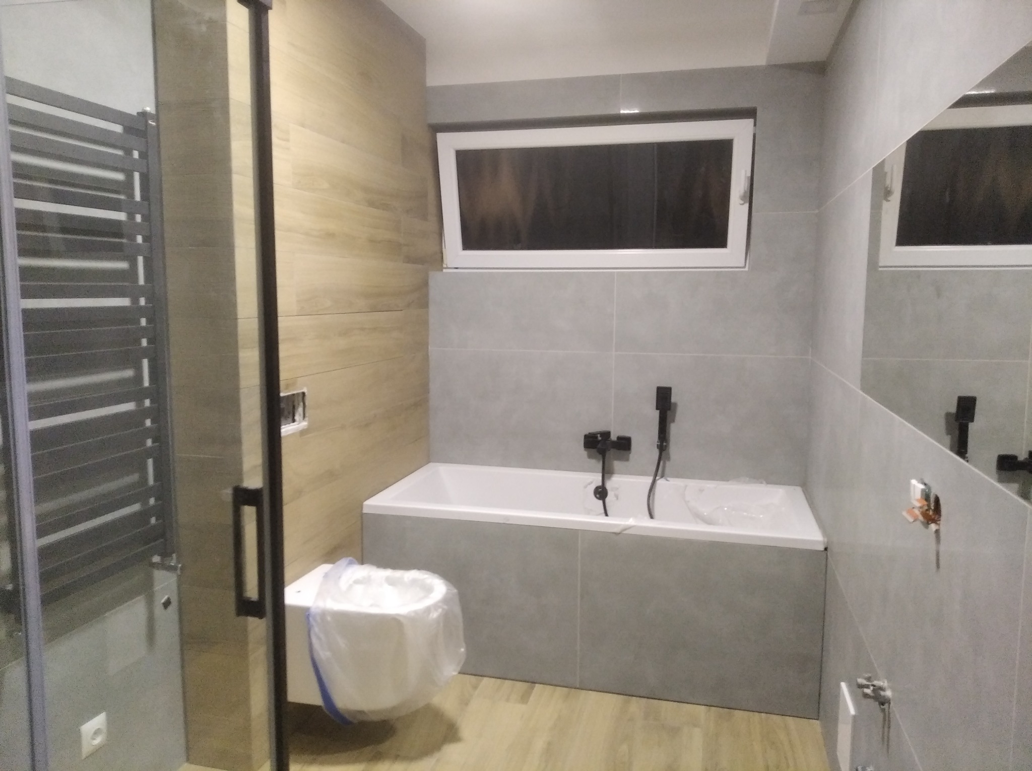 Nowoczesna łazienka z wanną obudowaną szarą płytką, drewnopodobną okładziną ścienną, grzejnikiem drabinkowym i ukrytym oknem, po remoncie, przed montażem armatury.