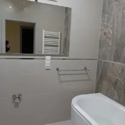 Wyremontowana łazienka z białą wanną, jasnymi płytkami i marmurową ścianą, widoczne lustro z odbiciem ręki trzymającej telefon i grzejnik drabinkowy.