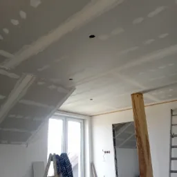 Sufit z płyt kartonowo-gipsowych w trakcie montażu i szpachlowania, widoczne łączenia płyt oraz otwory na oświetlenie, drabina z ubraniem roboczym i narzędzia w tle.