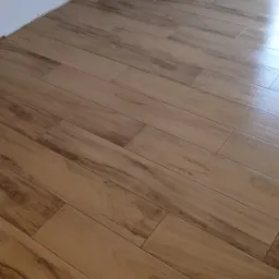 Ułożona pod kątem brązowa podłoga z płytek imitujących drewno, widoczna biała ściana z niedokończonymi gniazdkami elektrycznymi.