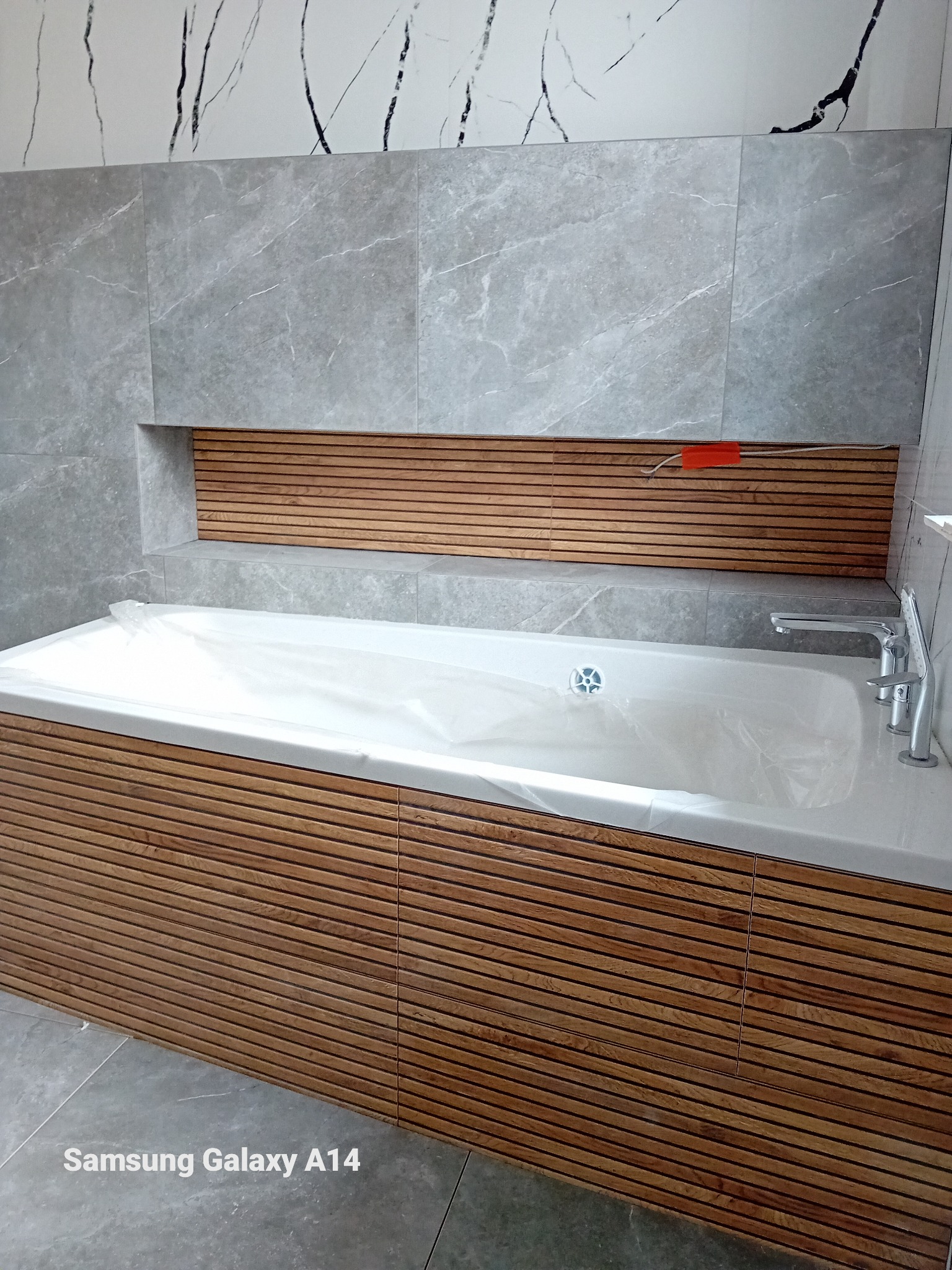 Nowoczesna wanna obudowana poziomymi panelami drewnianymi, zabezpieczona folią ochronną, z baterią umywalkową i odpływem, na tle szarych płytek imitujących marmur z czarnymi żyłami i wnęki...