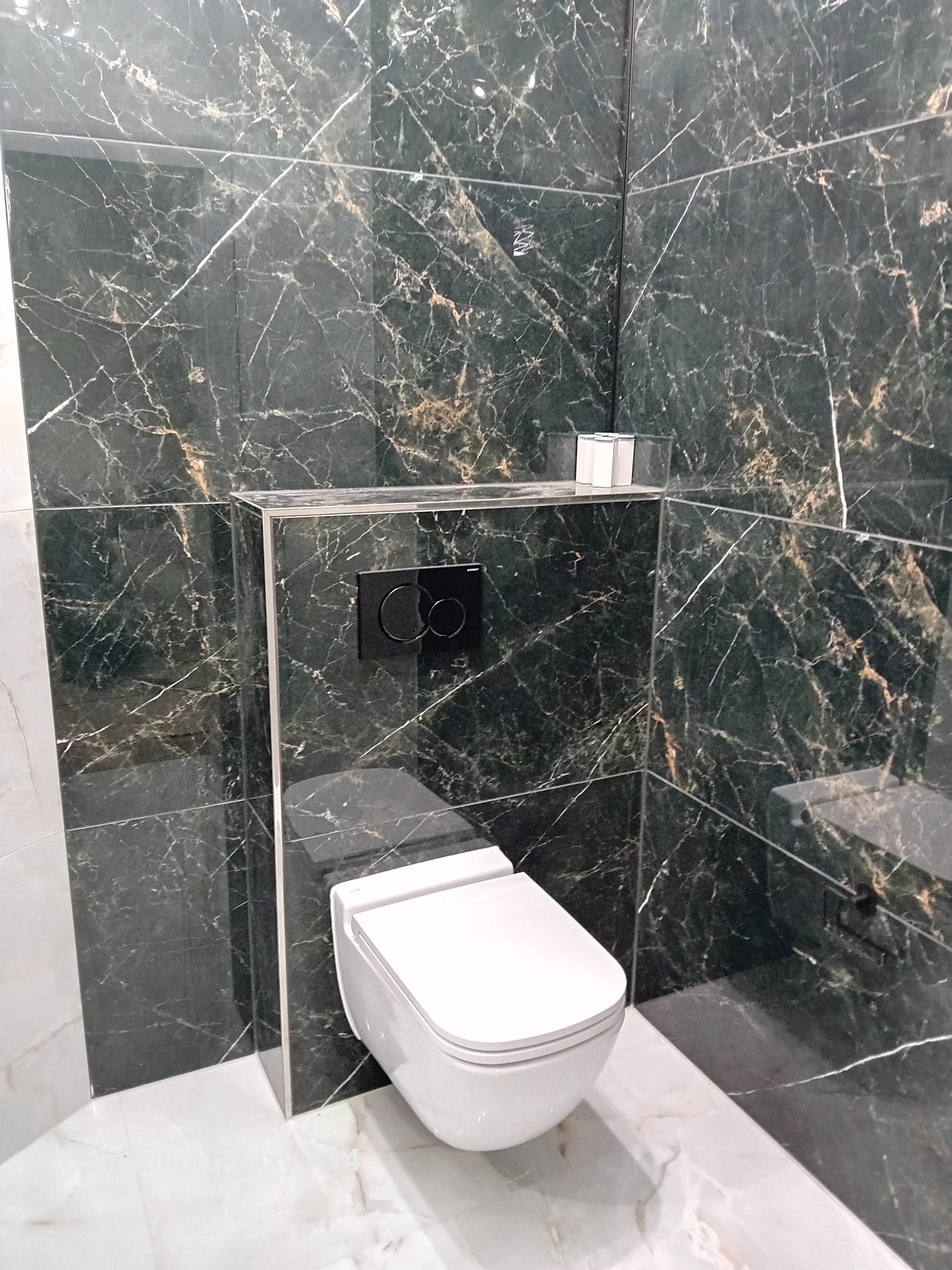Wisząca toaleta z czarnym przyciskiem spłukiwania na tle ściany wyłożonej ciemnozielonym marmurem z białymi żyłami, podłoga z jasnego marmuru.