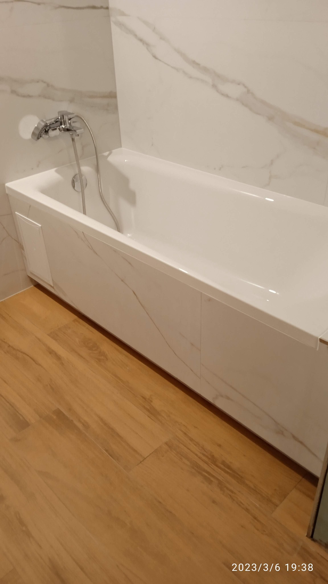 Biała wanna obudowana panelami imitującymi marmur, dopasowana do marmurowych płytek ściennych, z chromowaną baterią i drewnopodobną podłogą.