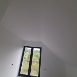 Karol Matuszewski - Poddasze ze świeżo pomalowanymi na biało ścianami i sufitem, widoczne otwory na gniazdka elektryczne i okno z widokiem na zieleń.