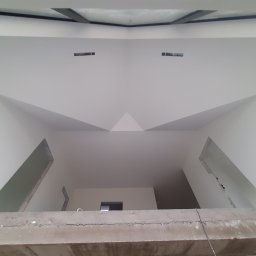 Karol Matuszewski - Surowe wnętrze z widocznymi ścianami po szpachlowaniu i malowaniu na biało, z fragmentem betonowej konstrukcji na pierwszym planie, widok z dołu na sufit z otworami na oświetlenie.