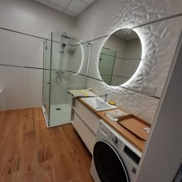 Karol Matuszewski - Nowoczesna łazienka z kabiną prysznicową, umywalką w zabudowie z pralką i okrągłym lustrem z podświetleniem LED na ścianie z trójwymiarową teksturą.