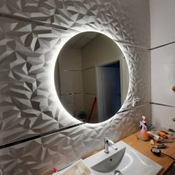 Karol Matuszewski - Łazienka po renowacji: ściana wyłożona trójwymiarowymi płytkami ceramicznymi w odcieniach szarości, okrągłe lustro podświetlane LED, biała umywalka z chromowaną baterią, widoczne narzędzia...