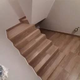 Schody wyłożone panelami imitującymi drewno, widok z góry, częściowo widoczna ściana z gniazdkiem elektrycznym i opakowanie materiałów budowlanych.