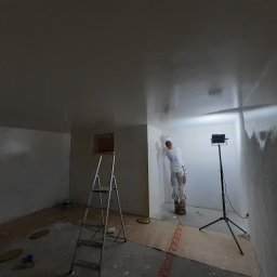 Karol Matuszewski - Mężczyzna szpachluje ścianę w pokoju, stojąc na podwyższeniu, z widoczną drabiną, lampą oświetleniową i narzędziami malarskimi.