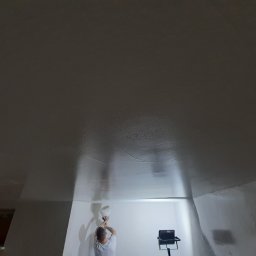 Karol Matuszewski - Praca na wysokości: mężczyzna szpachluje sufit w pomieszczeniu oświetlonym halogenem, widoczna faktura świeżo nałożonej masy.