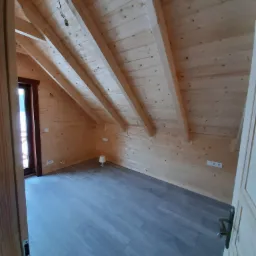 Poddasze wykończone drewnem z szarymi panelami podłogowymi, widok zza uchylonych drzwi, widoczne skosy, gniazdka elektryczne i minimalistyczna lampka.