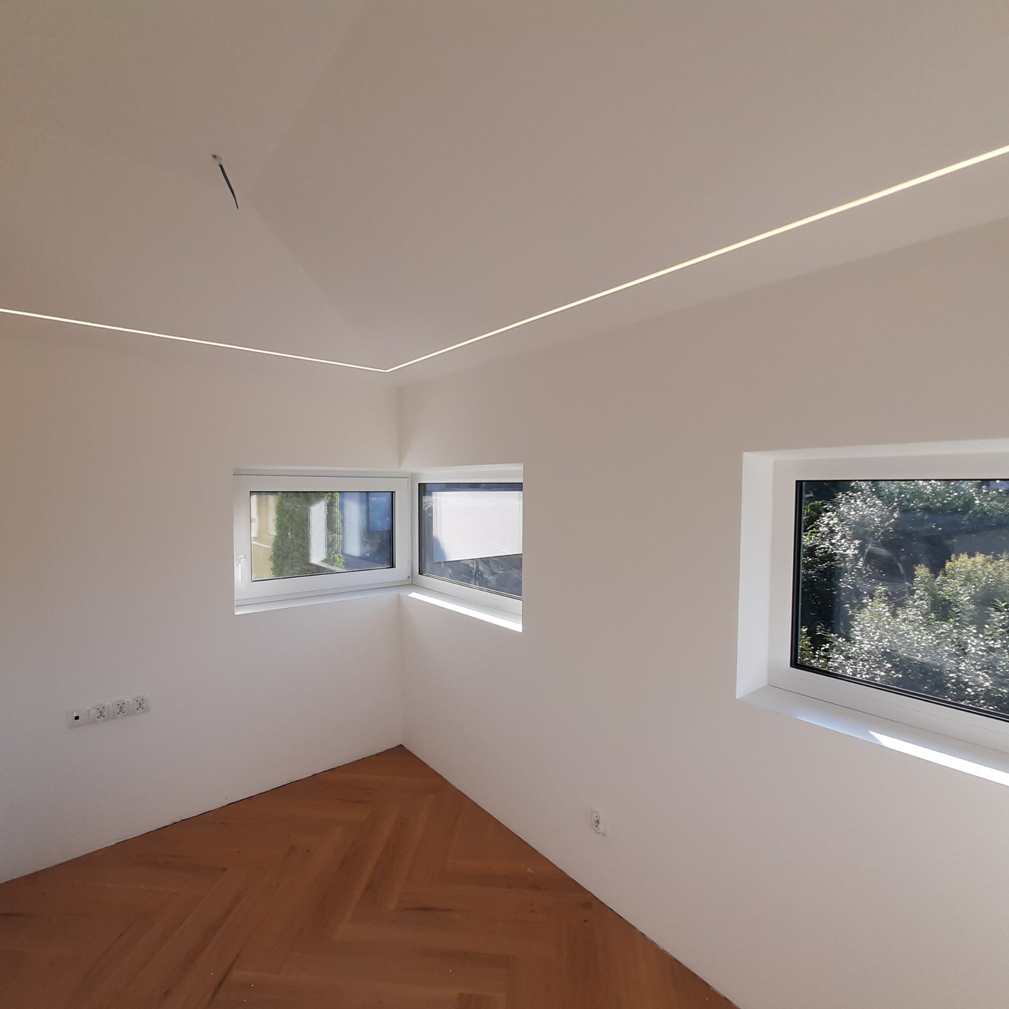 Wnętrze pokoju po malowaniu ścian na biało, z trzema oknami i drewnianą podłogą ułożoną w jodełkę oraz z oświetleniem LED w suficie.